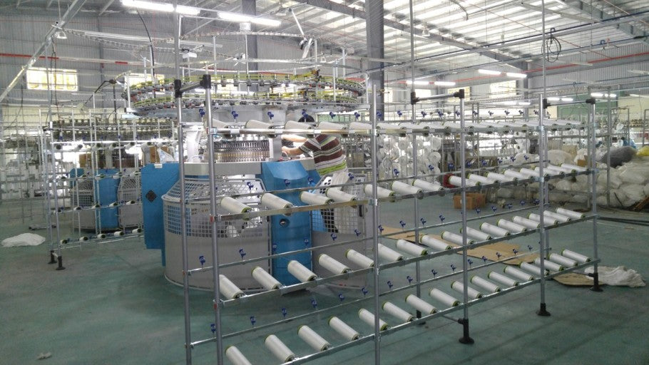 A large knitting machine in a factory is surrounded by racks of white yarn.