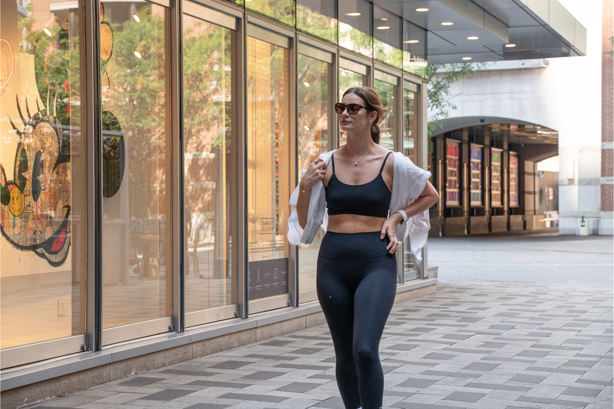 A young woman wearing sunglasses and a Orso Activewear SeaButter™ bra and leggings walks along a brick-paved sidewalk in a city shopping area, with a white shirt draped over her shoulders.